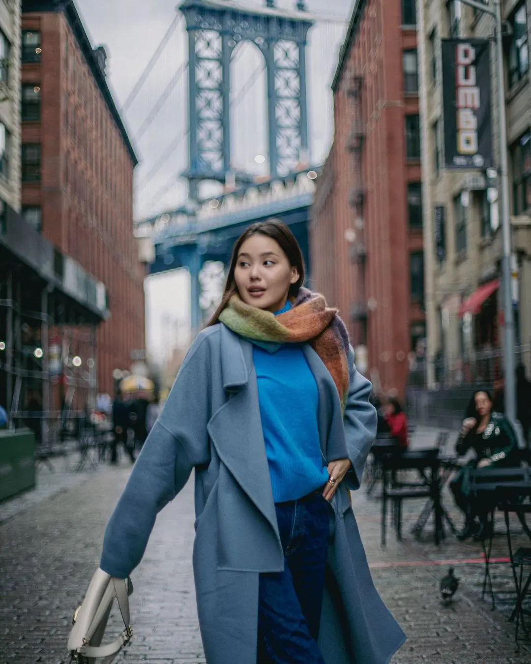 A street-style color photograph by Daykin Dayanch. A woman in a blue coat and a multi-colored scarf walks on a cobblestone street in DUMBO, Brooklyn. In the background, the blue-painted Manhattan Bridge is framed between red brick buildings, one of which has a vertical 'DUMBO' sign.