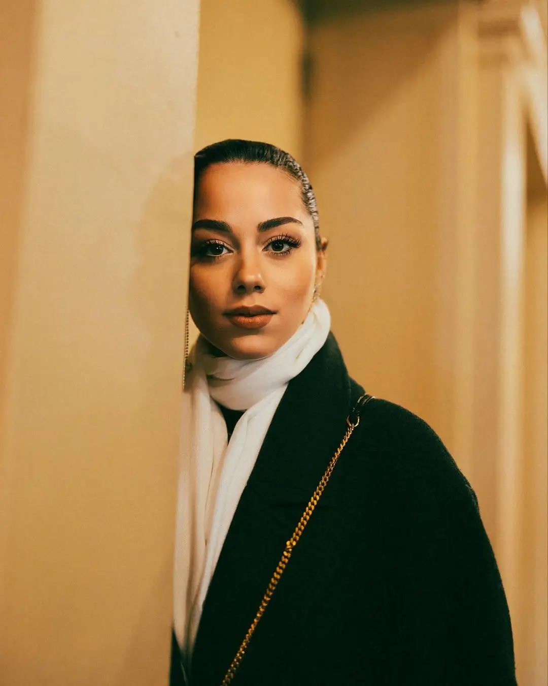 An elegant, warmly lit color portrait by Daykin Dayanch. A woman with her hair slicked back neatly looks out from behind a corner wall with a direct gaze. She is dressed in a dark coat and a soft white scarf, with the gold chain of a shoulder bag visible.