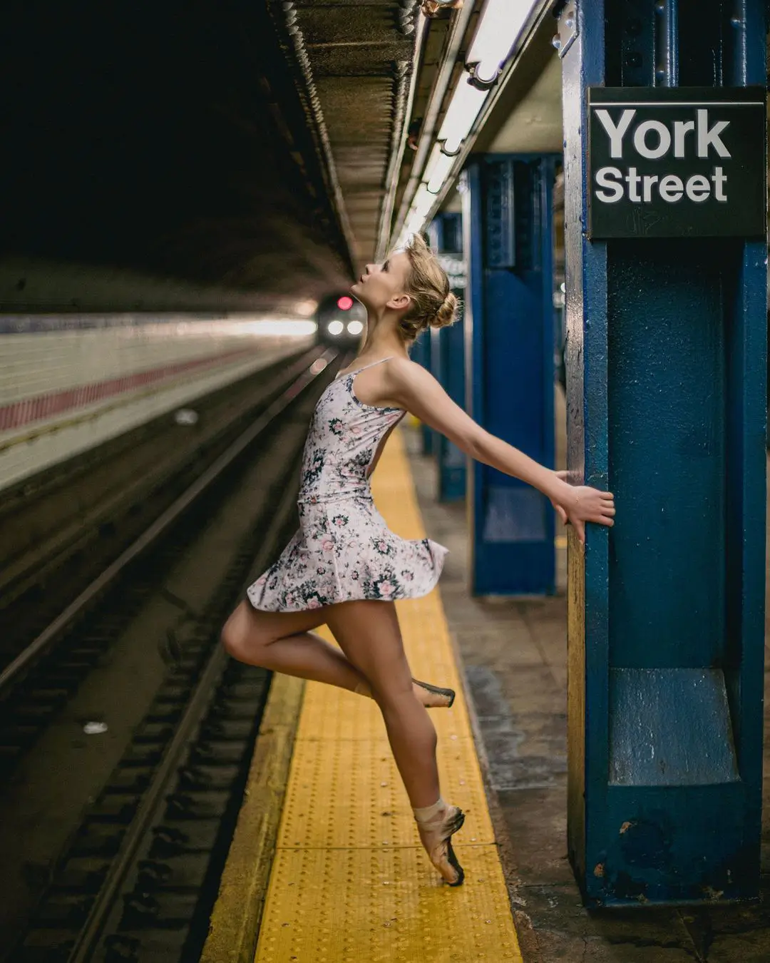 A striking color photograph by Daykin Dayanch, capturing the contrast between art and environment. A ballet dancer in a patterned leotard is frozen mid-leap, en pointe, on the yellow warning strip of a subway platform. She looks up as the lights of an approaching train emerge from the tunnel. A sign for the "York Street" station is mounted on a blue pillar beside her.
