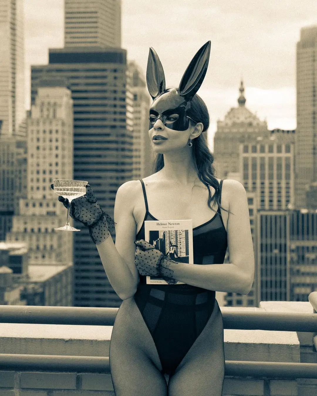A stylized, sepia-toned photograph by Daykin Dayanch. On a city rooftop, a woman poses in a black leotard, sheer polka-dot gloves, and a black mask with tall bunny ears. She looks off to the side while holding a coupe glass in one hand and a book by 'Helmut Newton' in the other. A dense cityscape of skyscrapers forms the background.