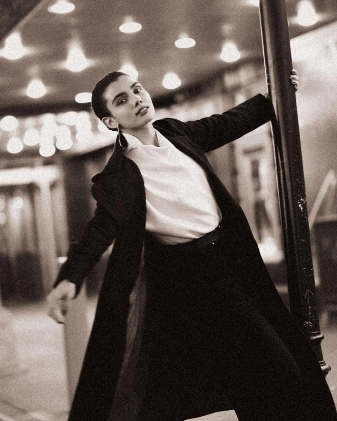 A chic, black-and-white photograph by Daykin Dayanch. A woman with her hair pulled back tightly, wearing a long dark coat over a white shirt, leans back against a pole in what appears to be a subway station. She looks confidently at the camera, with tiled walls and station lights blurred in the background.