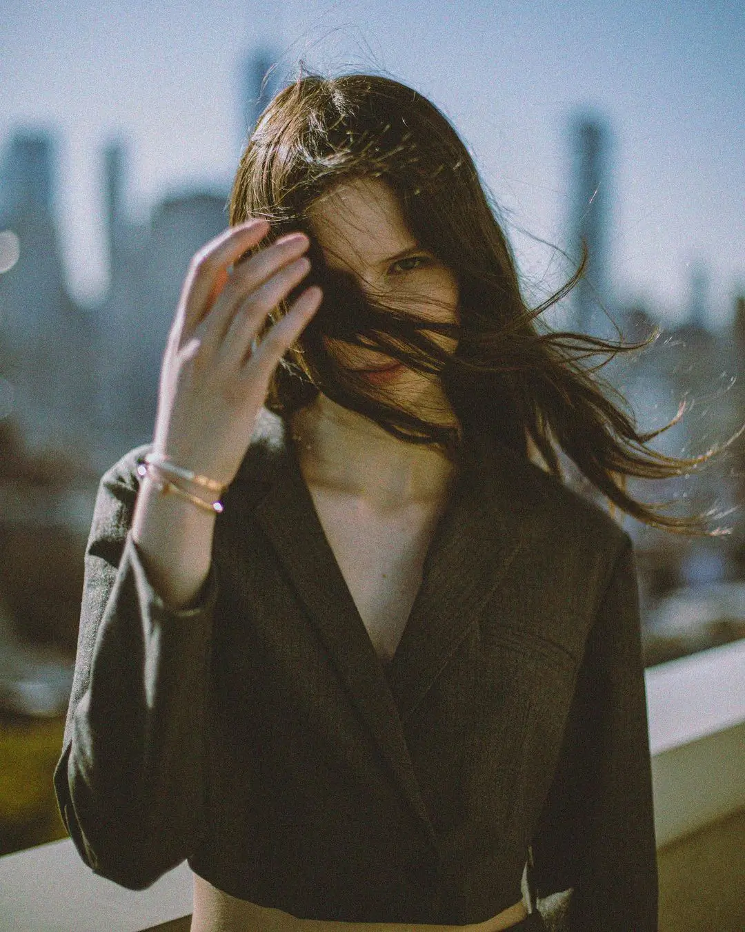A warm, film-style color photograph by Daykin Dayanch. A woman with long, dark hair stands on a sunny rooftop, looking at the camera from behind her windblown hair. She has one hand raised towards her face. In the background, a city skyline is softly blurred.