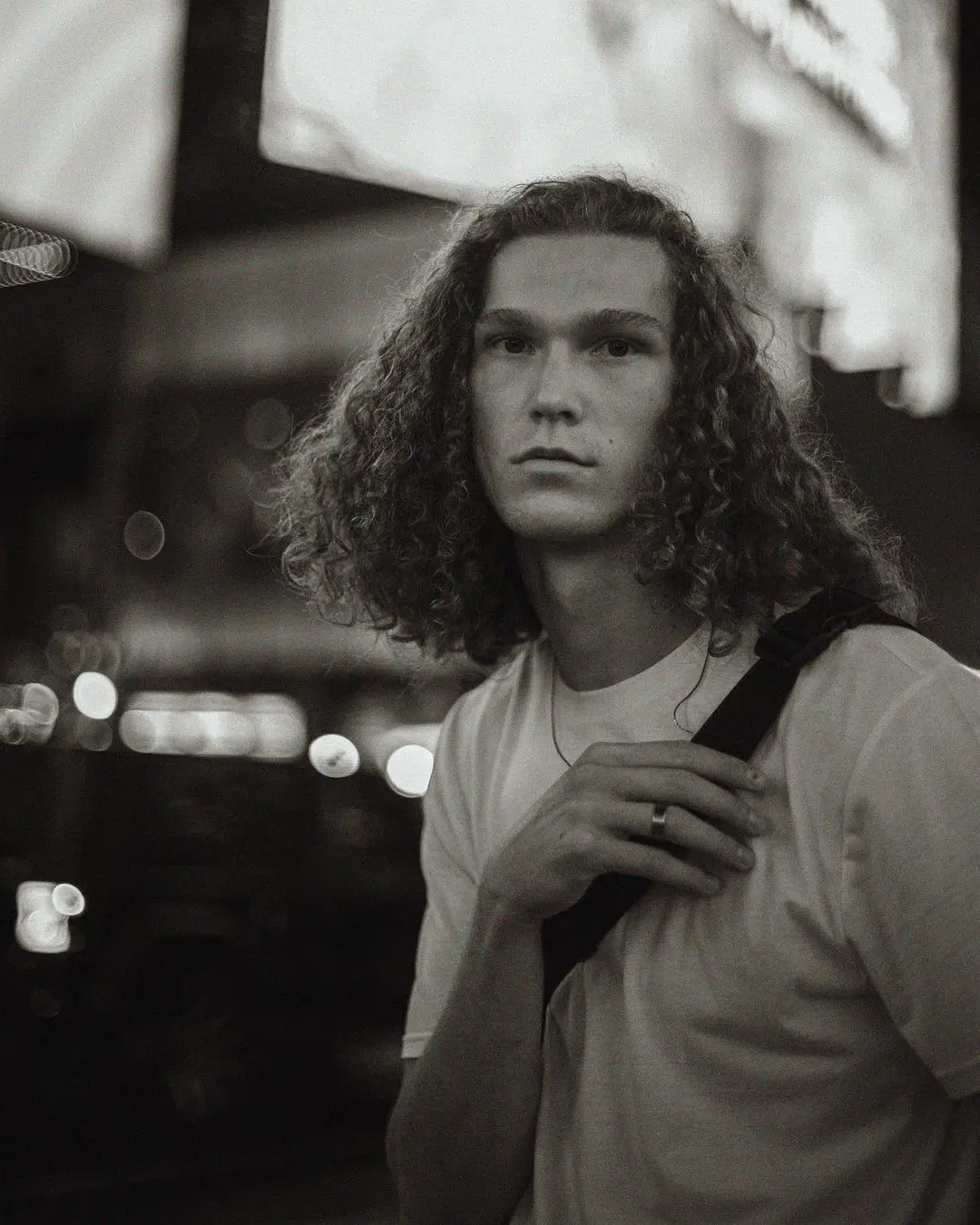 A black-and-white portrait by Daykin Dayanch, taken at night. A young man with long, curly hair, wearing a white t-shirt and a shoulder bag, looks directly at the camera. The background is soft and out of focus, showing blurred city lights and signs.
