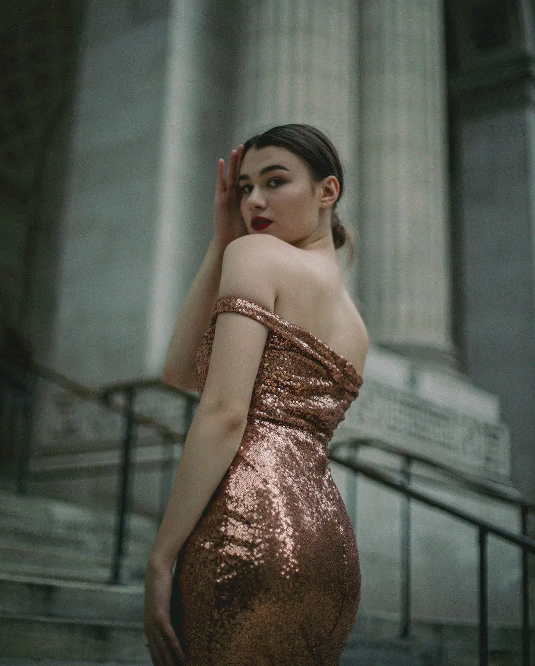 An elegant, film-style color photograph by Daykin Dayanch. A woman in a glamorous, off-the-shoulder gown made of rose-gold sequins poses on a grand stone staircase. She looks over her shoulder at the camera with a confident expression, her hand gently touching her temple. In the background, massive, fluted stone columns suggest a neoclassical building.