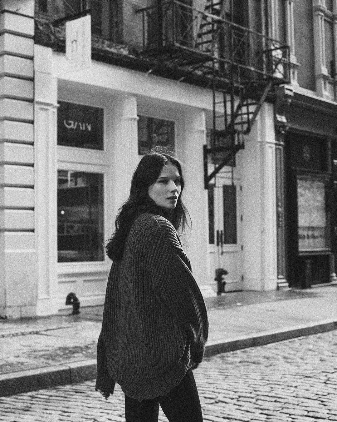 A black-and-white street photograph by Daykin Dayanch. A woman with long, dark hair, wearing an oversized, chunky-knit sweater, looks back over her shoulder as she walks down a cobblestone street. The background features classic city buildings, one of which has an external fire escape.