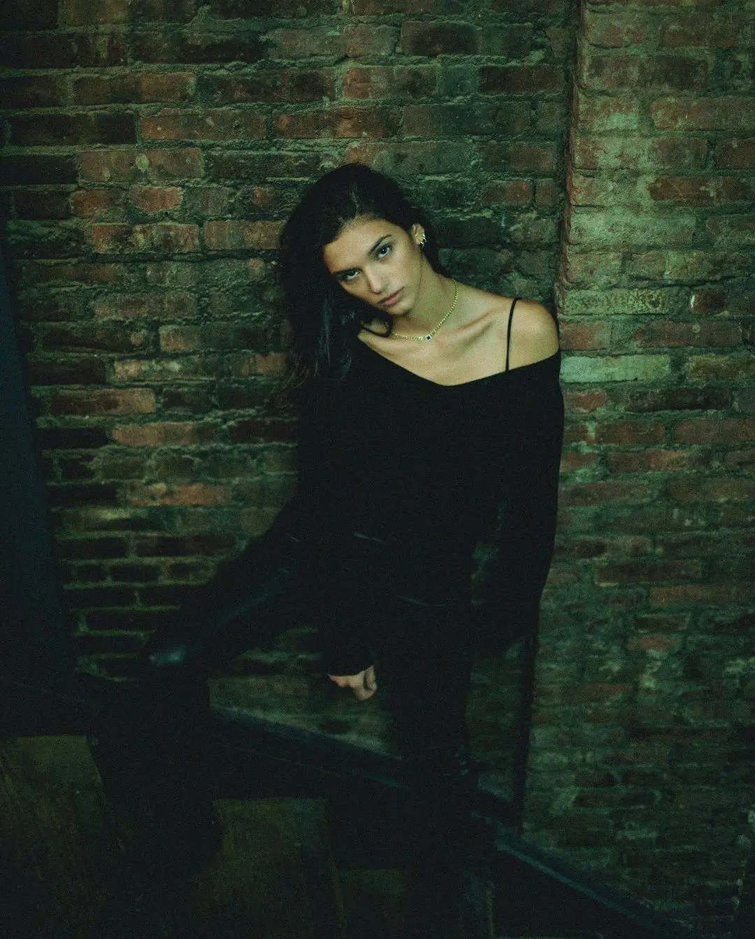 A high-angle, moody color portrait. A woman in a black top and a necklace with an eye-shaped pendant leans against an exposed brick wall in a dimly lit space. She looks up at the camera with a direct gaze, her face highlighted by a single light source.