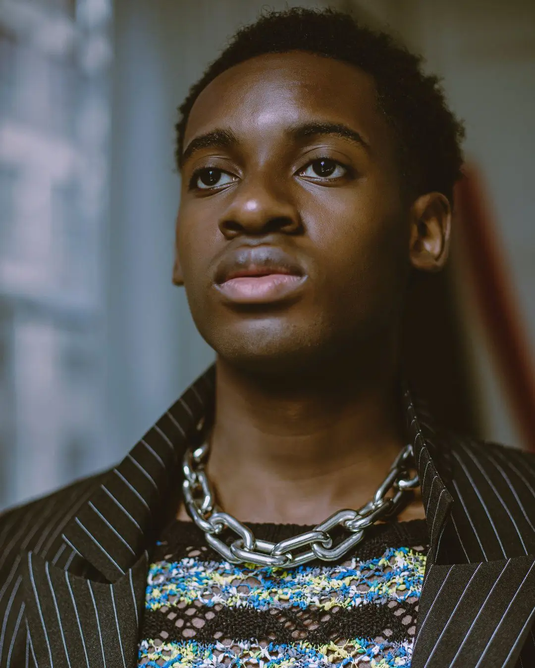 A close-up, editorial-style color portrait by Daykin Dayanch. A young Black man looks up and away with a thoughtful expression. He is wearing a colorful textured sweater, a thick, silver-colored chain-link necklace, and a pinstriped blazer. The background is a softly blurred industrial or studio space