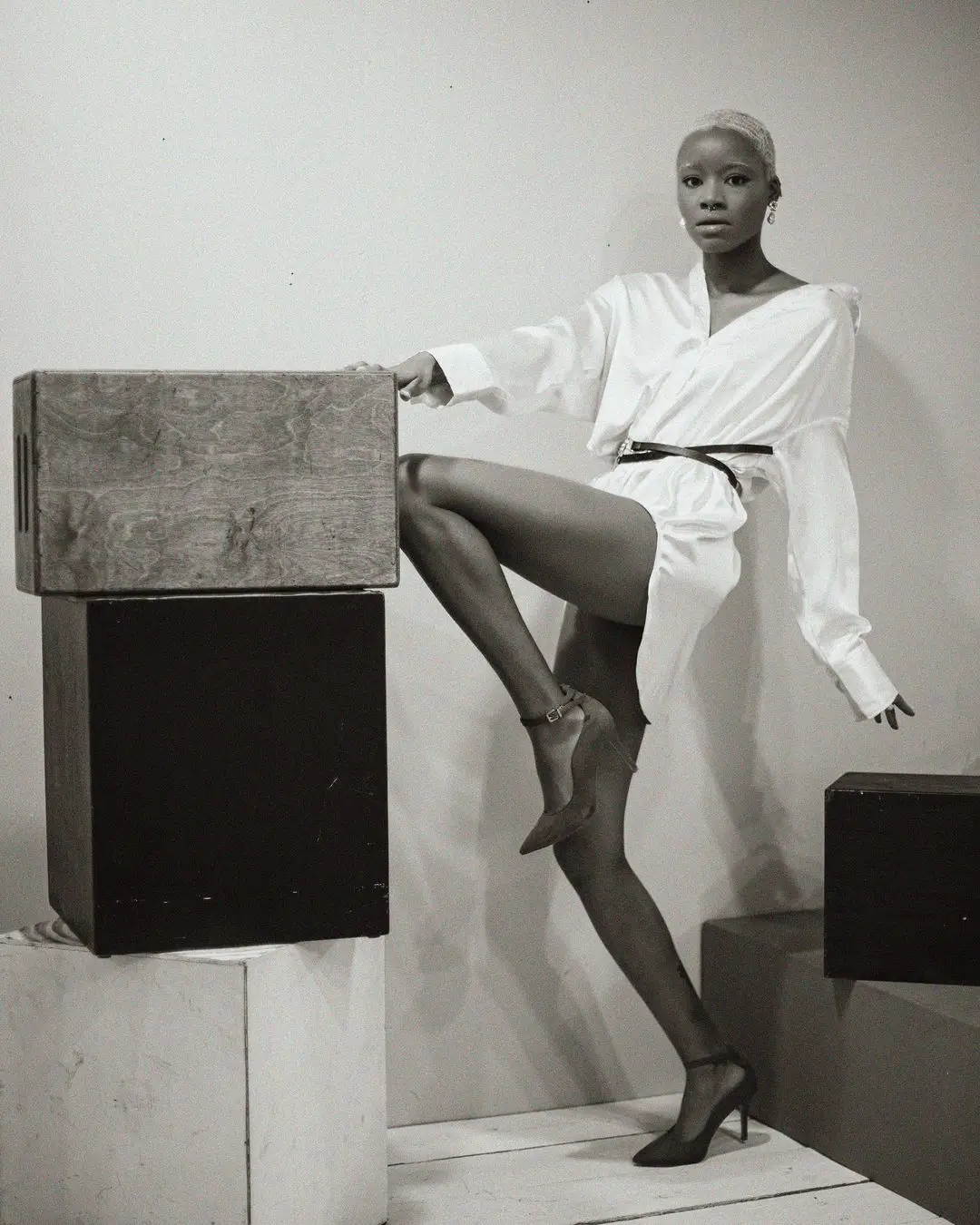 A full-length, black-and-white studio photograph by Daykin Dayanch. A woman with very short, light-colored hair strikes a powerful pose, resting a raised leg on a stack of geometric blocks. She wears a belted, oversized white shirt and stiletto heels, and looks directly at the camera with a strong gaze.