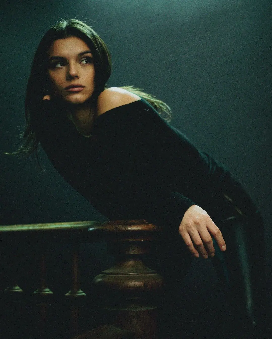 A moody, film-style color portrait. A woman in a black, wide-necked top leans forward over a wooden banister. She looks up and to the side thoughtfully, her face illuminated by an overhead light in a dark room.