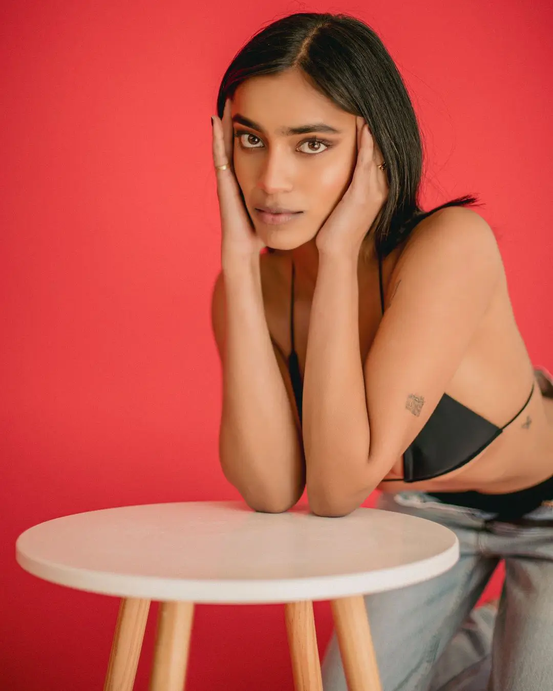 A photograph by Daykin Dayanch. A woman with straight black hair poses confidently in front of a bold red background. She leans on a round white stool, wearing light denim jeans and a black halter-style top. Her hands frame her face, highlighting her intense gaze and natural makeup. Small tattoos are visible on her upper arm and side.