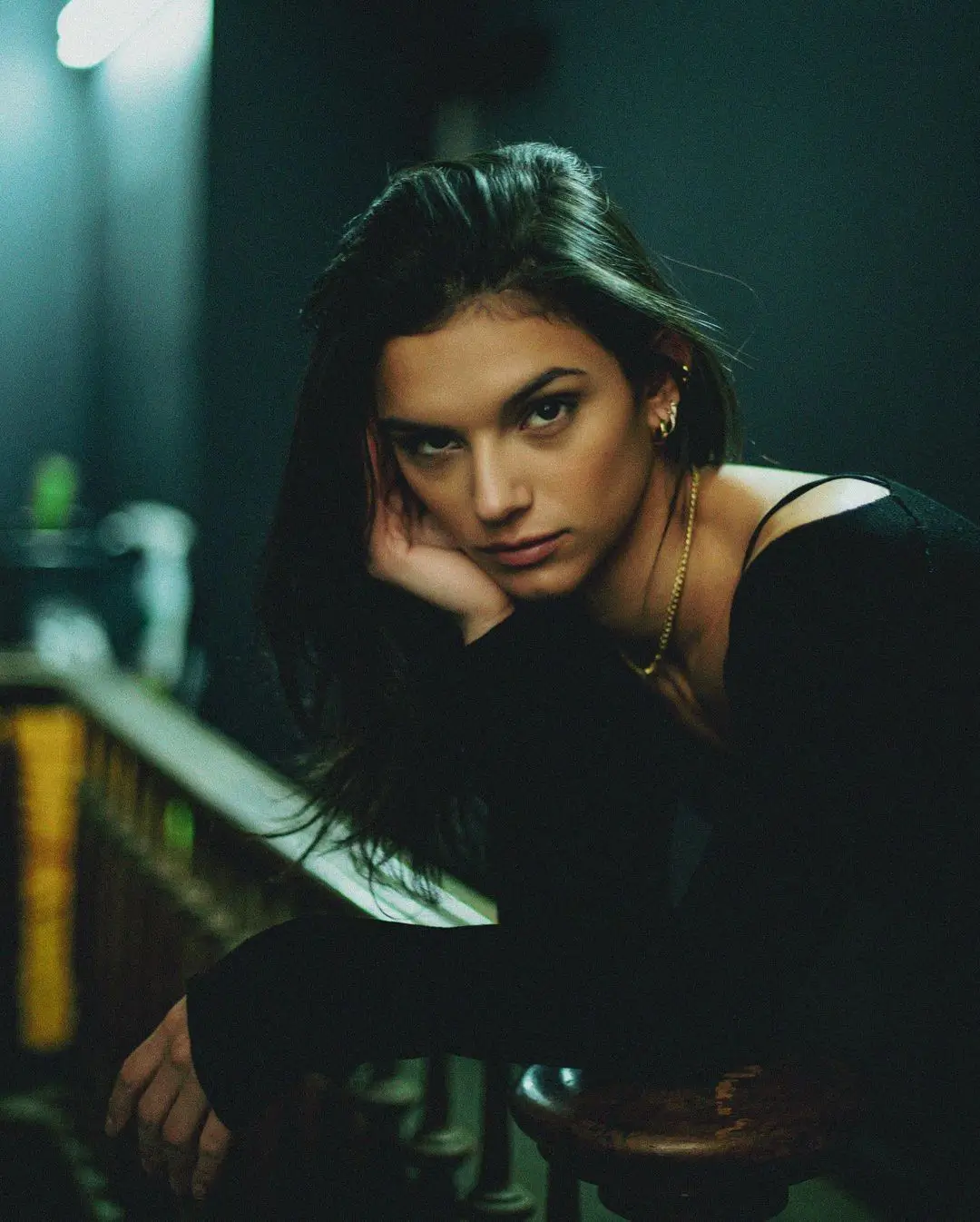A moody, film-style color portrait. A woman with long, dark hair rests her cheek on her hand as she leans against a banister, looking intently at the camera. She wears a black long-sleeved top and a gold necklace, illuminated by a soft light in a dimly lit room.