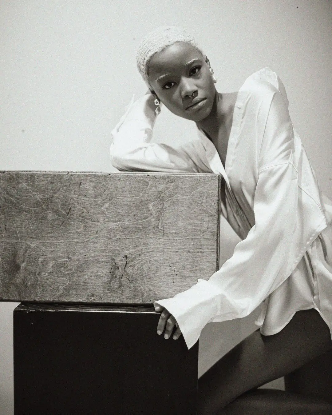 A black-and-white studio photograph by Daykin Dayanch. A woman with very short, light-colored hair leans her elbow on a rustic wooden box and looks directly at the camera. She is wearing a white satin shirt and has several piercings in her ears and nose.