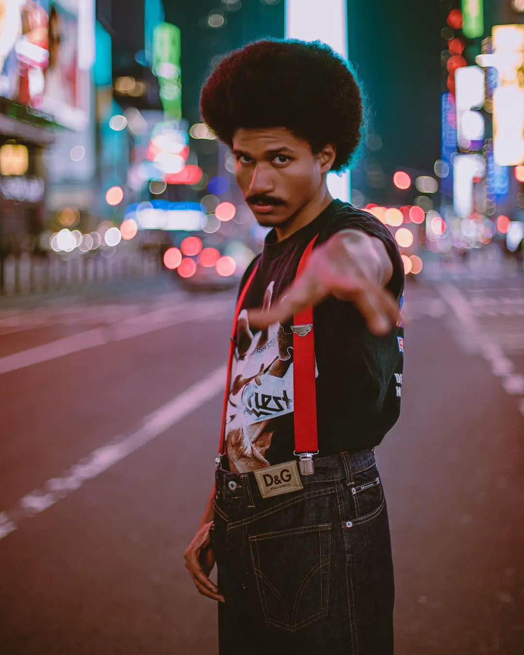 A vibrant, street-style color portrait by Daykin Dayanch. A man with a large afro and a mustache stands in the middle of a city street at night, pointing directly at the camera. He wears a graphic t-shirt, bright red suspenders, and jeans with a D&G logo. The background is filled with the red and white bokeh of city lights.