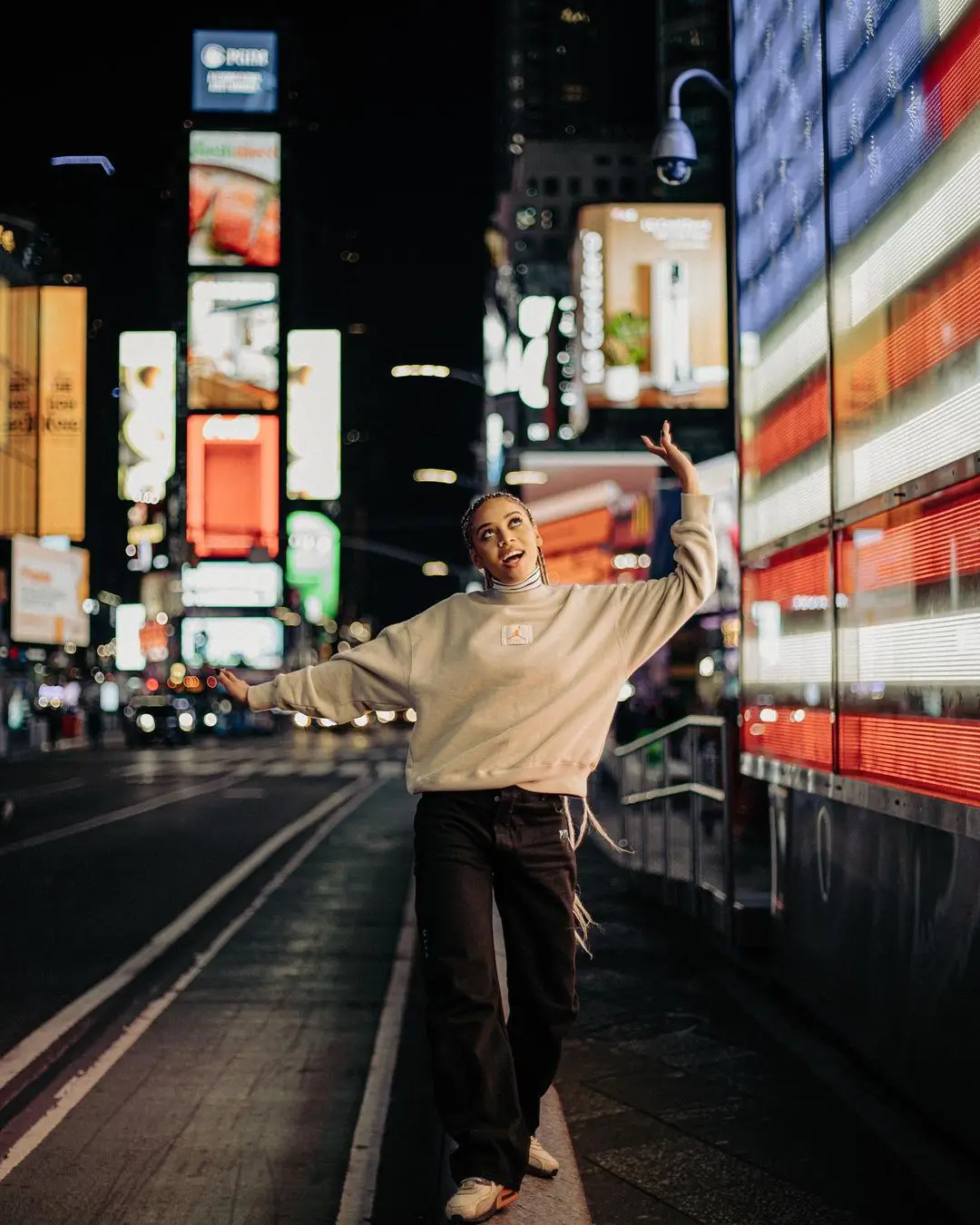 A joyous color photograph by Daykin Dayanch. A woman with long micro braids walks through Times Square at night with her arms spread wide, looking up in wonder. She is illuminated by the glow of a massive vertical LED screen to her right, which displays a pattern resembling an American flag.