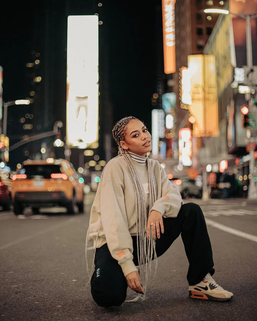 A street-style color photograph by Daykin Dayanch, taken at night. A woman with long, blonde and brown micro braids kneels on one knee in the middle of a city street, looking at the camera. She is wearing a beige sweatshirt, black pants, and Nike sneakers. The background is a blur of city life, including the bright lights of Times Square billboards and a yellow taxi cab