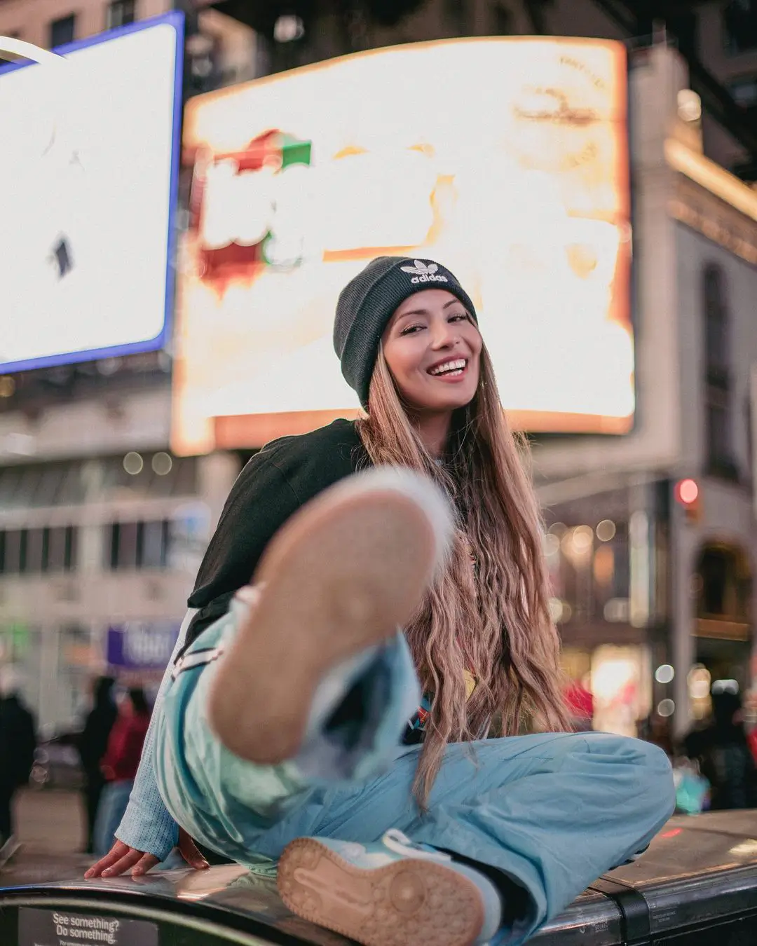 A playful, low-angle color photograph by Daykin Dayanch. A smiling woman with long hair, wearing an Adidas beanie, sits on a utility box in Times Square at night. She kicks one leg playfully towards the camera, so the sole of her sneaker is featured in the foreground. The background is a vibrant blur of massive, glowing billboards.