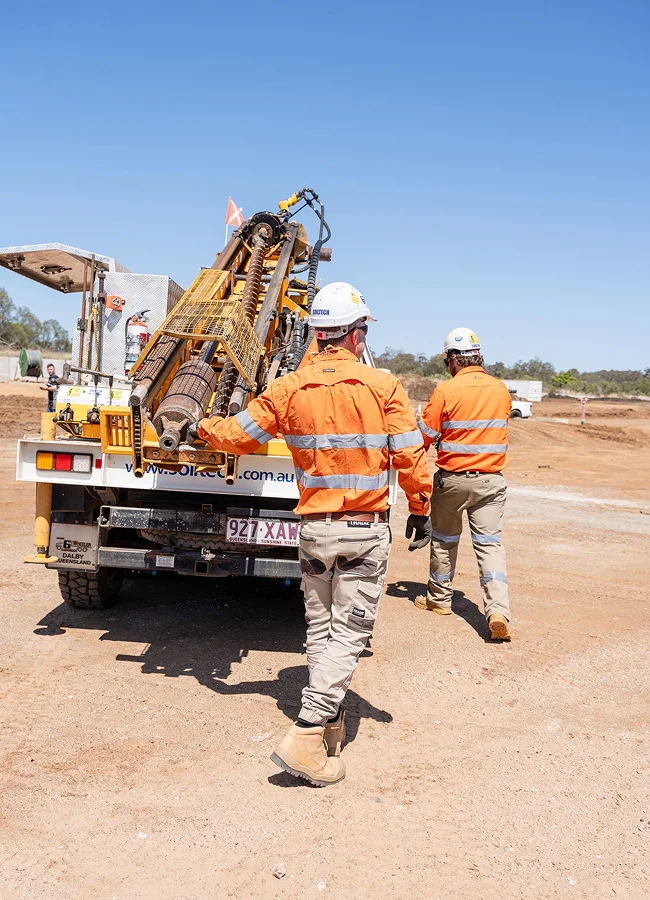 Two workers in orange safety jackets and helmets walking beside a truck carrying drilling equipment at a construction site under a clear blue sky.