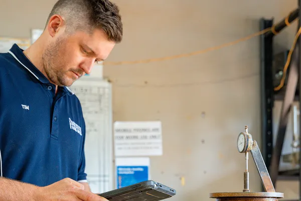 Man wearing a navy blue shirt labeled “Tim” using a tablet, with a measuring dial gauge on a nearby table.
