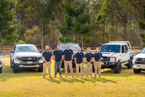 Six members of the Soilttech team standing in a line on grass outdoors, flanked by four white trucks, with trees and a fence in the background.