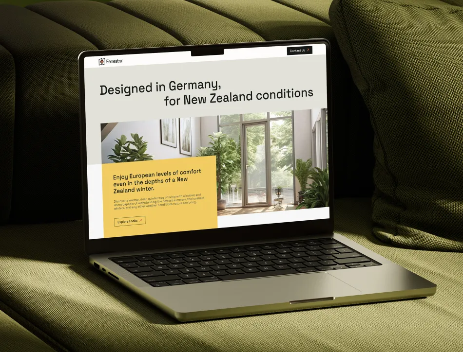 Laptop on olive green couch displaying a real estate or architecture website with text "Designed in Germany, for New Zealand conditions" and an image of a modern glass building