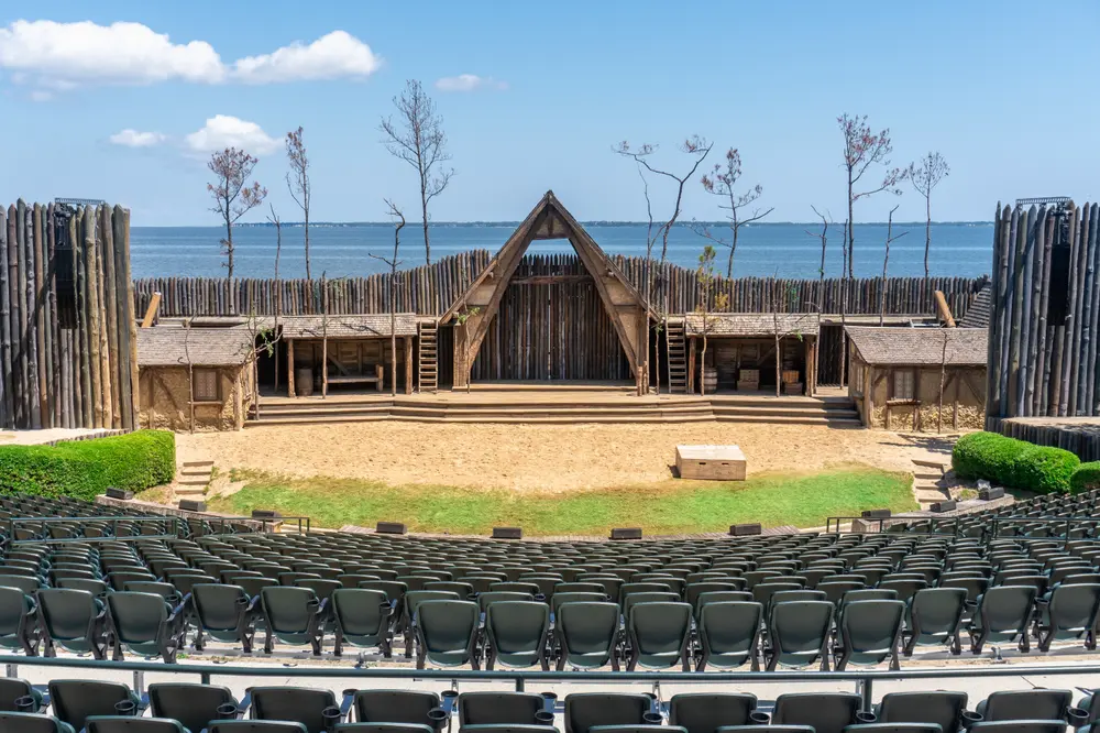 Outdoor theater seating facing a rustic wooden fort with clear skies and a water backdrop, representing The Lost Colony.
