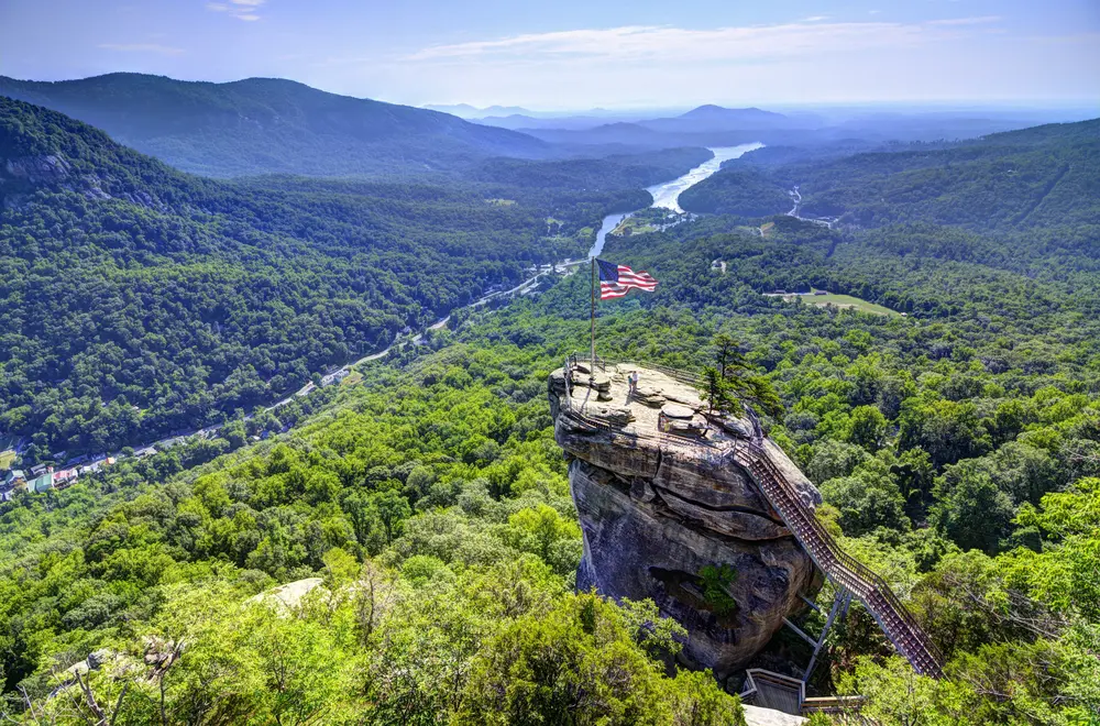 Overlooking Chimney Rock State Park with expansive green forest, winding river, and American flag atop rock formation.