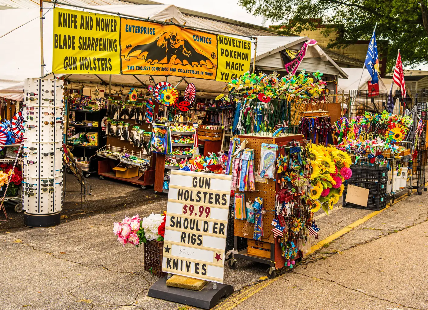 A vibrant market stall at The Raleigh Market selling gun holsters, shoulder rigs, knives, and a variety of colorful wind spinners and artificial flowers.