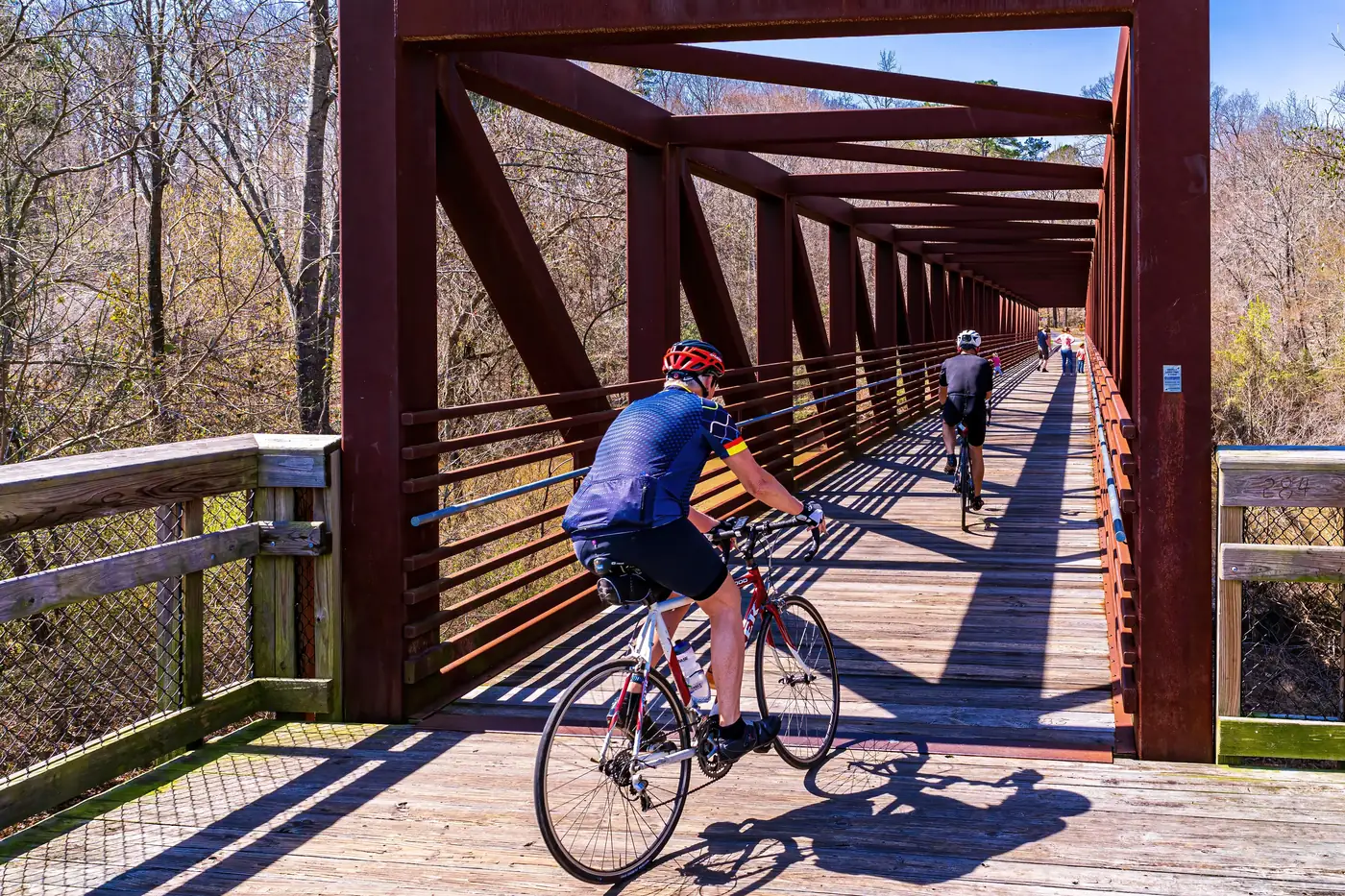 Cyclist and pedestrians on Neuse River Trail bridge with leafless trees in the background.