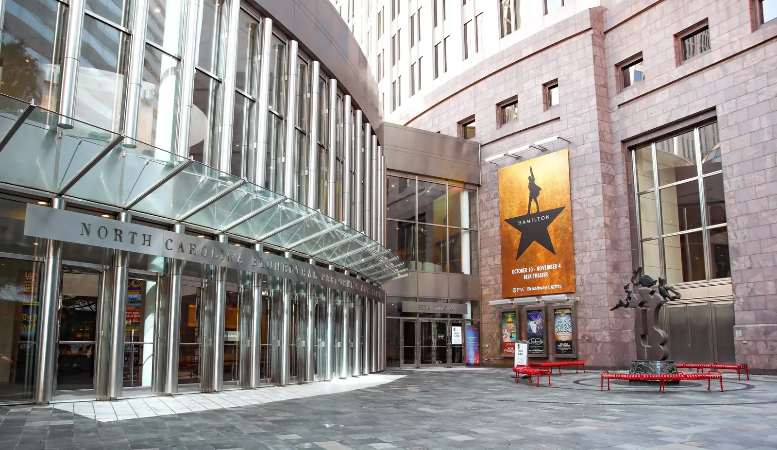 Exterior view of Blumenthal Performing Arts Center with glass facade, sculpture, and 'Hamilton' show banner.
