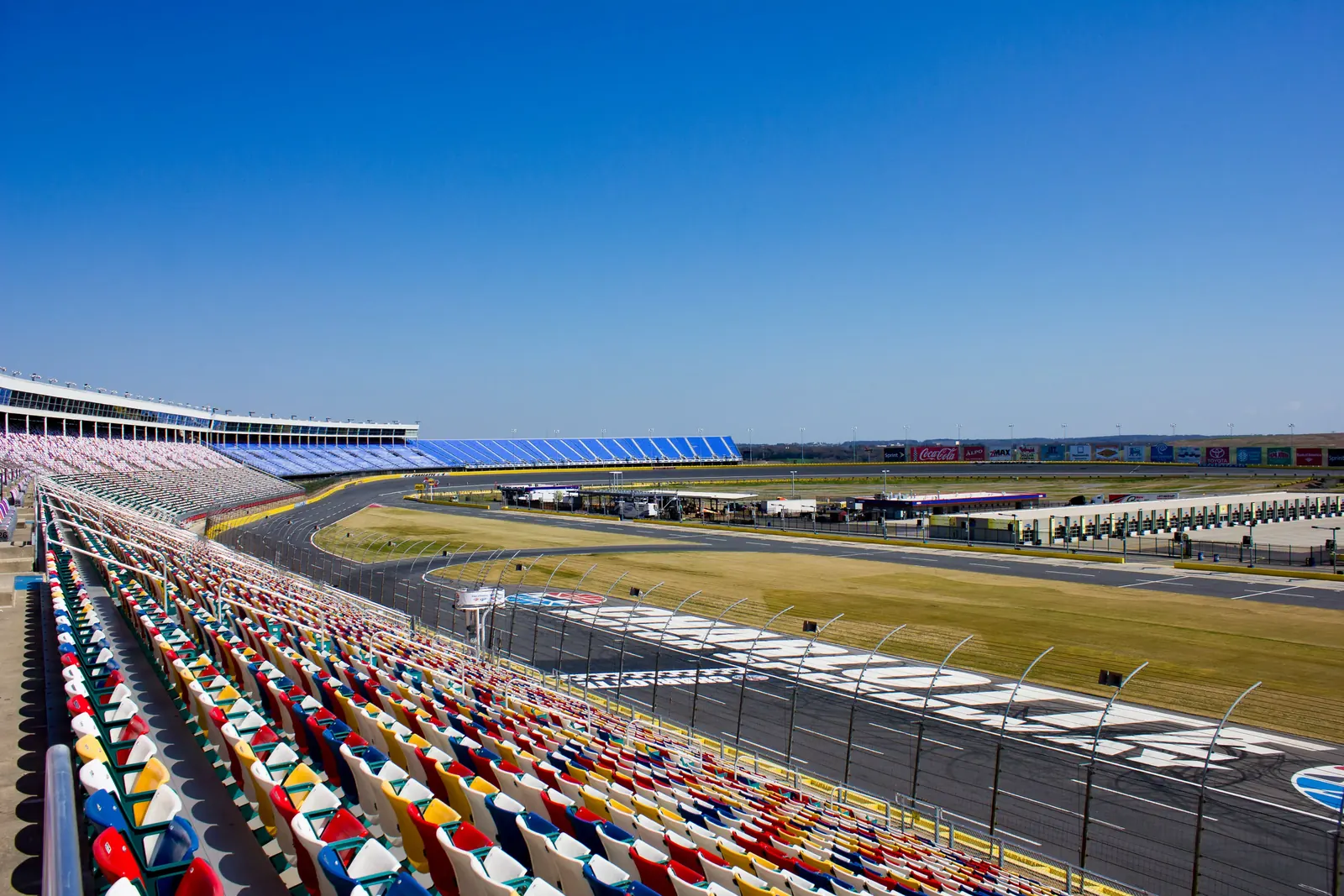 Sunny view of Charlotte Motor Speedway showing colorful grandstands and race track with clear blue sky.
