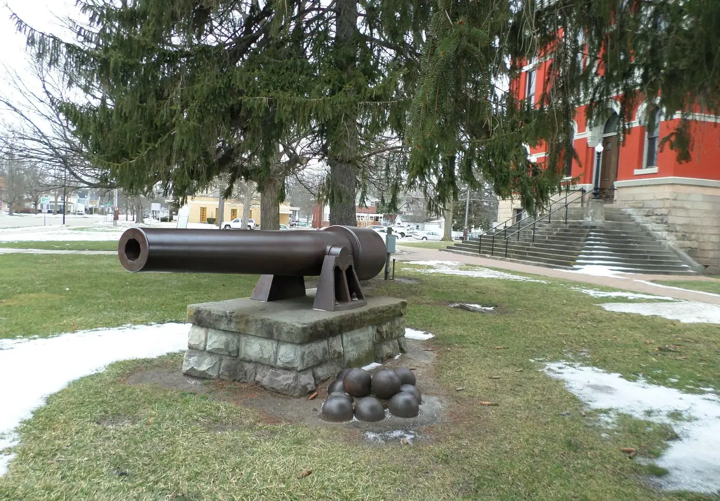 Antique cannon on display with cannonballs in front of the red Charlotte Museum of History building with steps and a green tree.