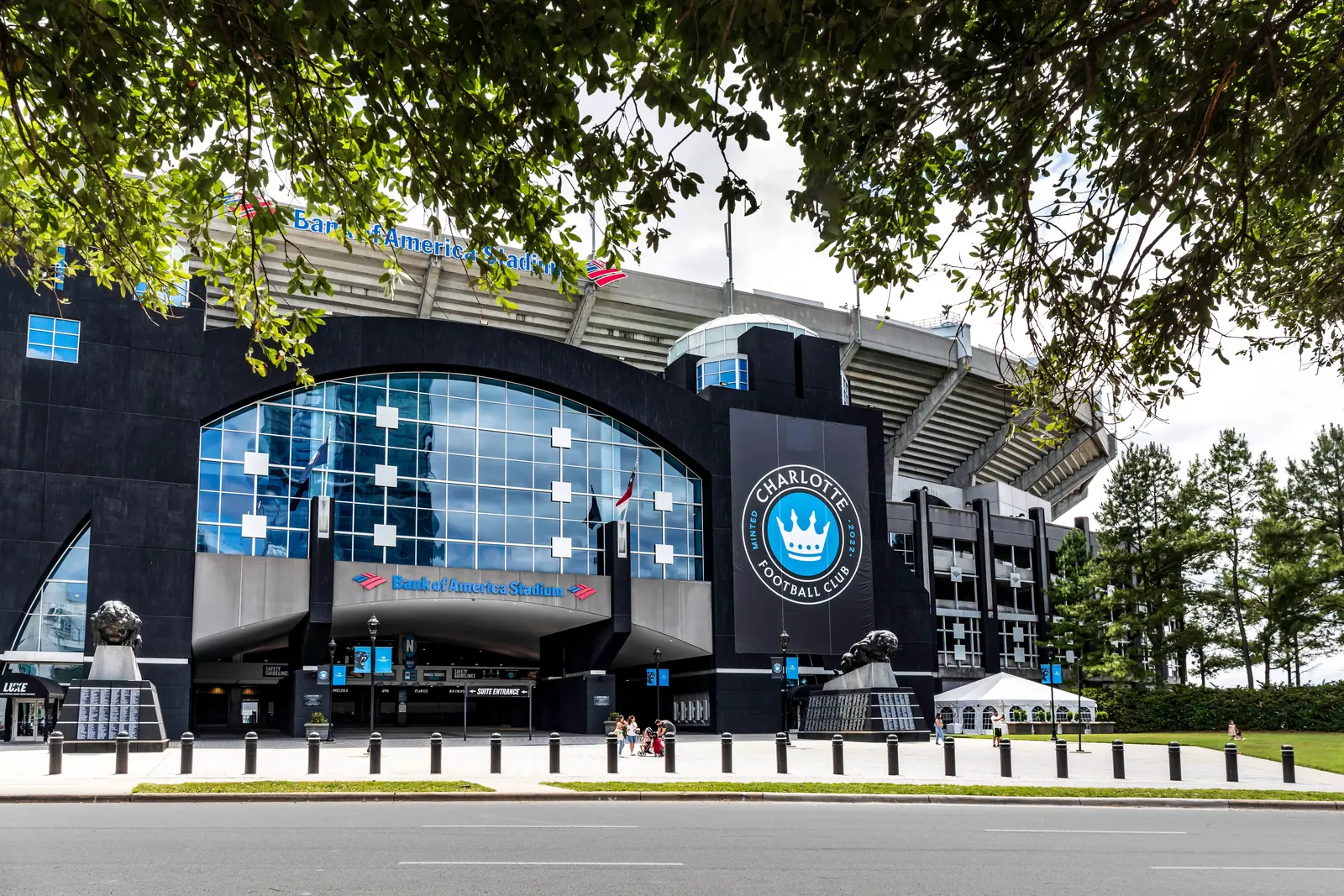 Front view of Bank of America Stadium with large glass windows, a statue, and the Charlotte Football Club logo.