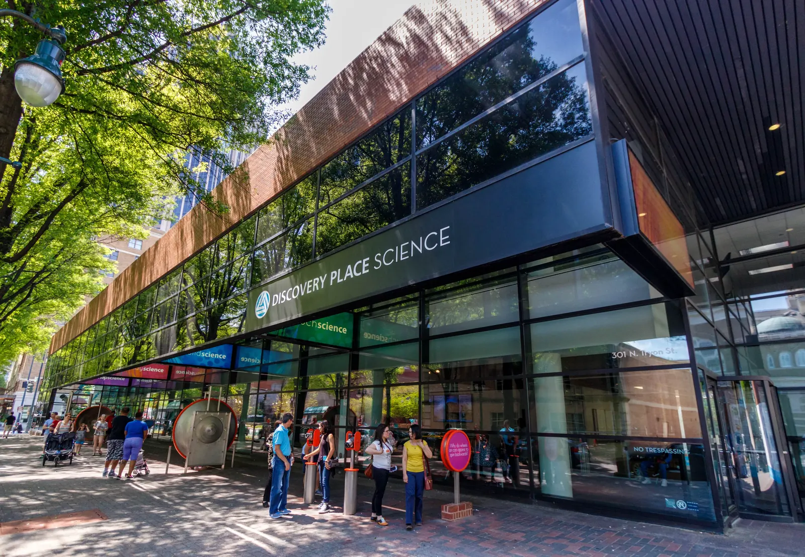 Exterior view of Discovery Place Science with visitors outside the entrance and reflective glass facade showcasing trees and buildings.