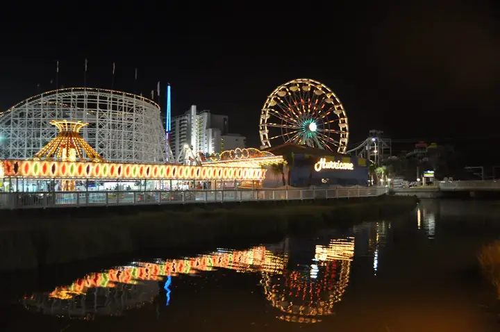 Night view of Family Kingdom Amusement Park with illuminated ferris wheel and roller coaster, reflected in water.