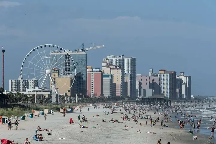 Sunny day at Myrtle Beach with people on the sandy shore and a Ferris wheel near tall buildings in the background.