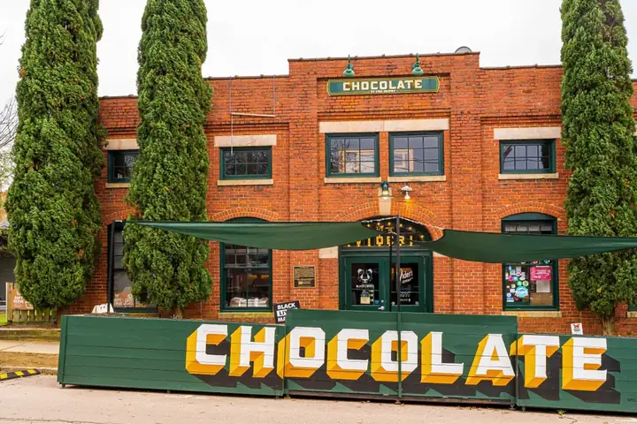 Brick building in The Warehouse District with "CHOCOLATE" sign, flanked by large greenery, and shaded entryway.