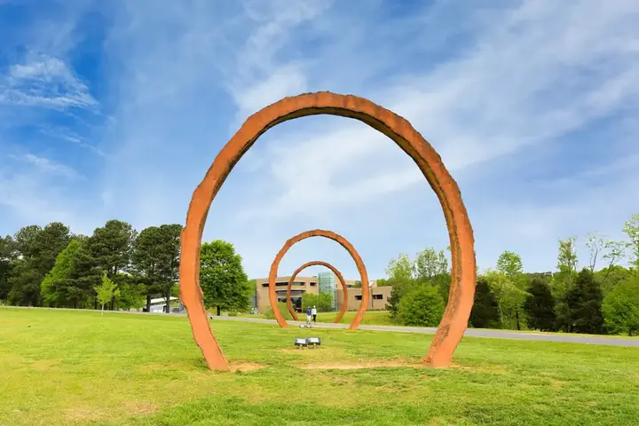Rusty large-scale arch sculpture on grass at North Carolina Museum of Art with blue sky and trees in background.