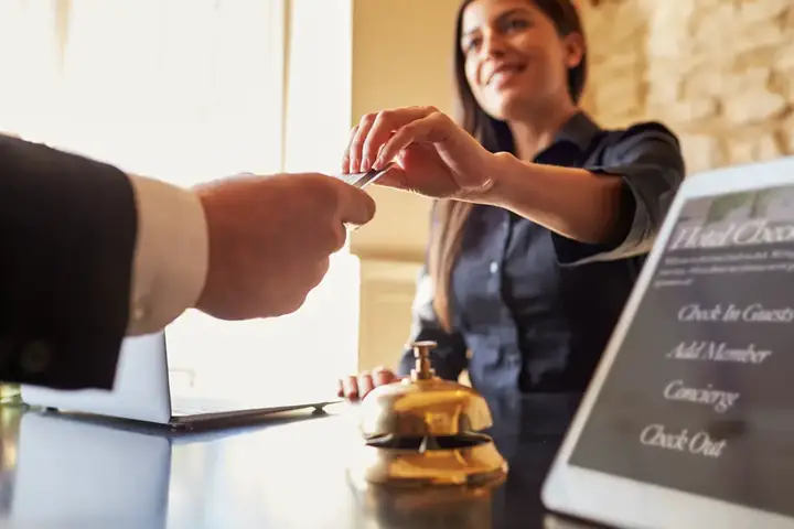 Hotel reception scene with smiling clerk handing key card to guest; tablet displaying 'Check In, Concierge, Check Out' services visible.