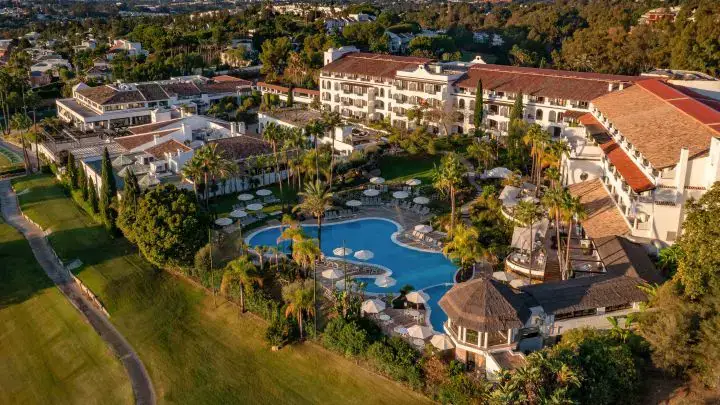 Aerial view of Dunes Village Resort showing white buildings, multiple swimming pools surrounded by palm trees, and adjacent golf course.