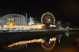 Night view of Family Kingdom Amusement Park with illuminated ferris wheel and roller coaster, reflected in water.