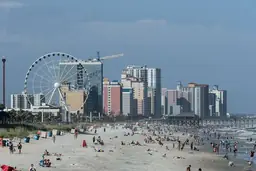 Sunny day at Myrtle Beach with people on the sandy shore and a Ferris wheel near tall buildings in the background.