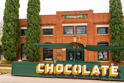 Brick building in The Warehouse District with "CHOCOLATE" sign, flanked by large greenery, and shaded entryway.