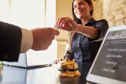 Hotel reception scene with smiling clerk handing key card to guest; tablet displaying 'Check In, Concierge, Check Out' services visible.