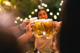 Friends clinking beer mugs together at Raleigh Beer Garden with glowing lights in the background.