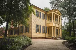 Colonial-style yellow house at Mordecai Historic Park with lush greenery and brick chimney.