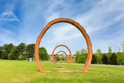 Rusty large-scale arch sculpture on grass at North Carolina Museum of Art with blue sky and trees in background.