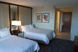 Well-lit hotel room with two queen beds, nightstand with lamp, artwork, and entrance door at Raleigh Marriott City Center.
