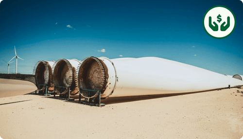 Refurbished wind turbine blades laid on ground with standing turbines in the background under clear blue sky.