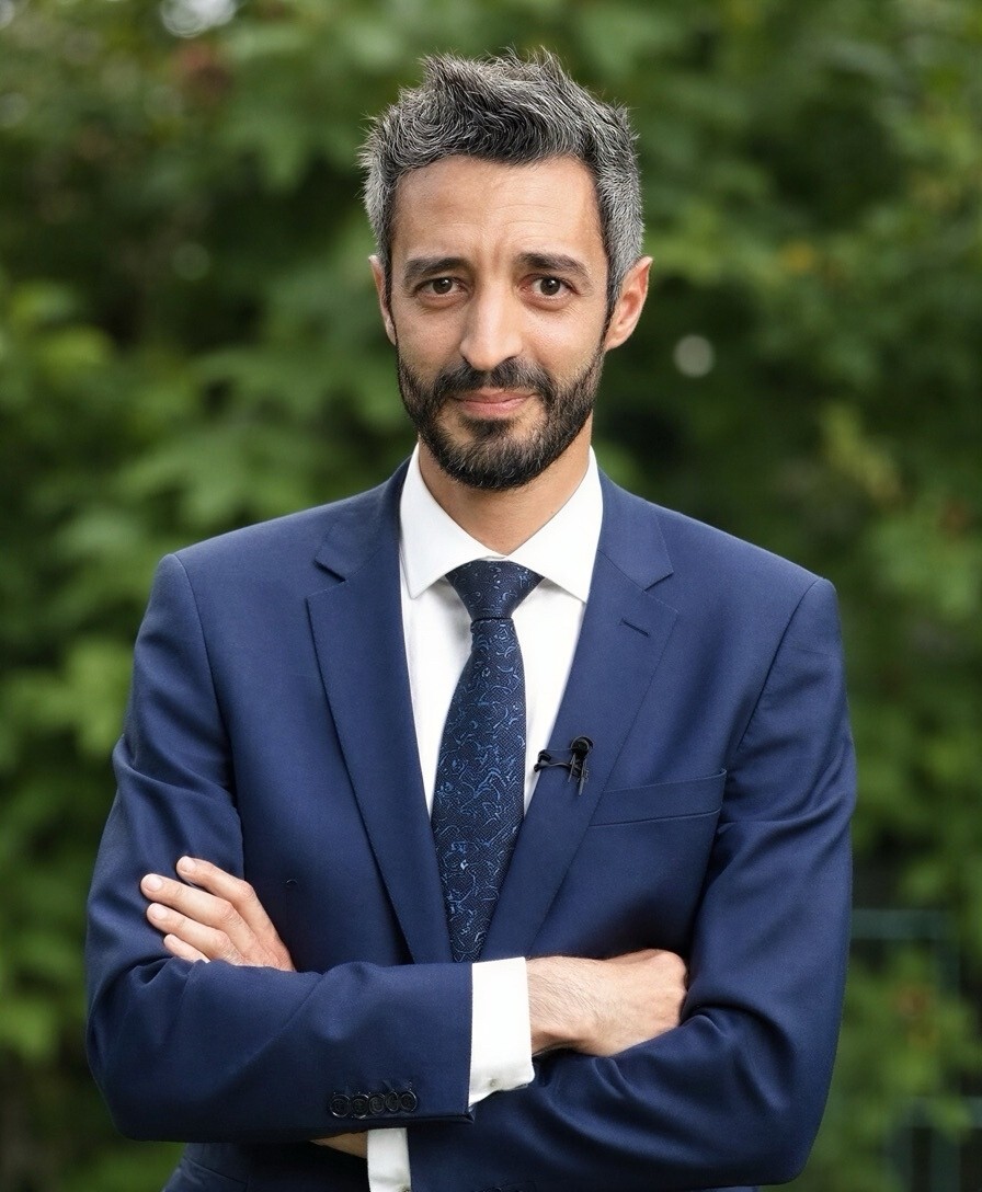 Man with gray hair and beard wearing blue suit and tie, standing with arms crossed outdoors.