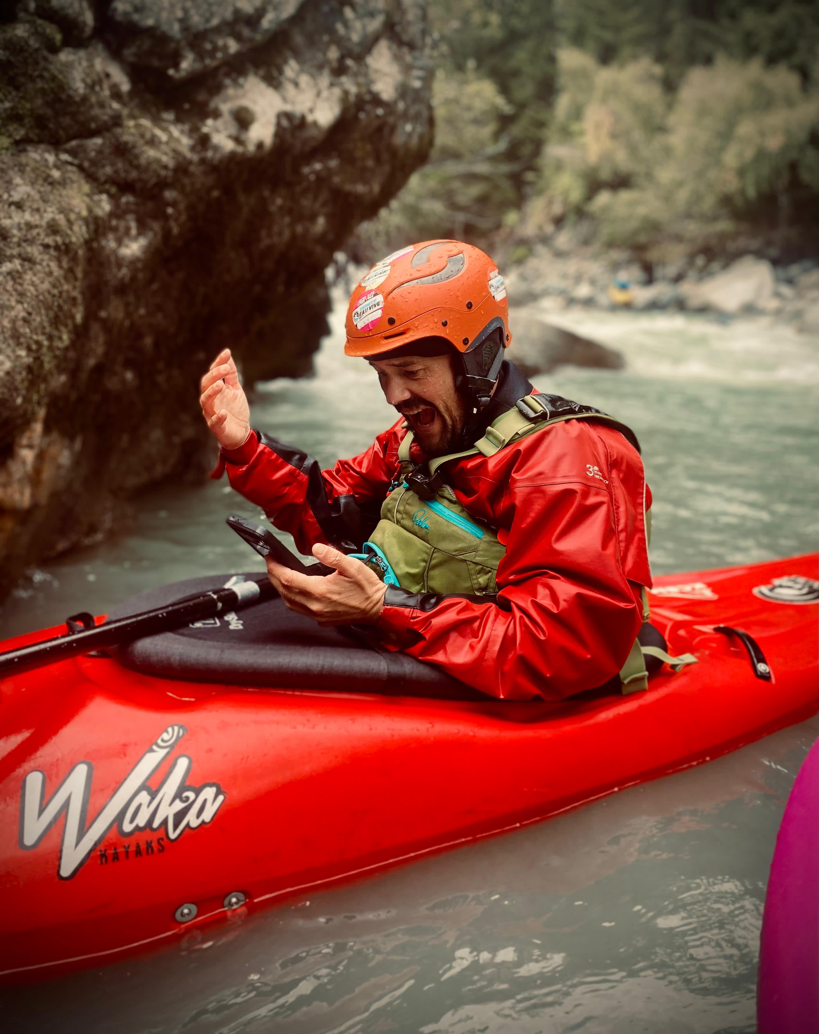 Man in orange helmet and red jacket sitting in a red Waka kayak holding a phone, frustrated